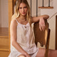 A woman seated on a wooden chair, wearing a white camisole and matching shorts with lace details.